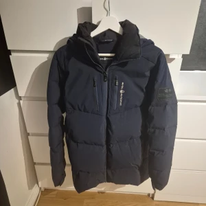 Mörkblå pufferjacka från Sail Racing - Säljer en mörkblå pufferjacka från Sail Racing med huva och dragkedja framtill. Jackan har två bröstfickor med dragkedja, logga på ärmen och snyggt mönstrad insida av huvan. Perfekt för kalla dagar och riktigt stilren look. Är som S i storlek