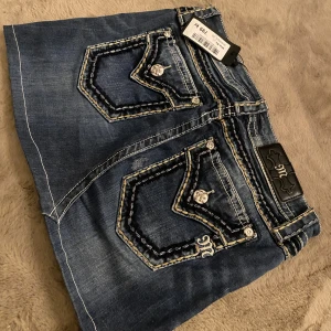 Jeanskjol från Miss Me, stl 29 - Snygg jeanskjol från Miss Me med coola kontrastsömmar och dekorativa fickor med silvriga knappar. Klassisk blå denim med broderade detaljer och logga på bakfickan. Perfekt för dig som gillar streetstyle och vill sticka ut lite extra. 