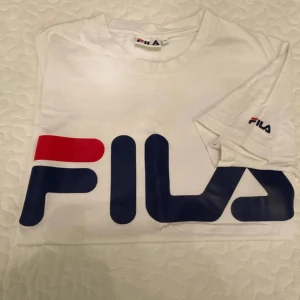 Vit FILA t-shirt med stor logga - Vit t-shirt från FILA i bomull med stor, ikonisk FILA-logga i blått och rött framtill. Klassisk rund hals och korta ärmar. Liten logga även på ärmen. Perfekt för en sportig och avslappnad stil.