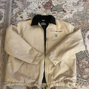 Beige Carhartt Detroit Jacket M - Säljer en beige Carhartt Detroit Jacket i storlek M. Jackan har svart krage i manchester och klassisk  Carhartt-logga på bröstet. Yttertyget är slitstarkt och jackan har dragkedja framtill samt flera fickor.