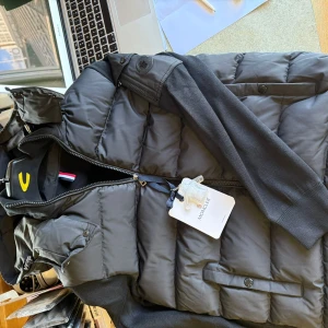 Svart pufferjacka från Moncler - Snygg svart pufferjacka från Moncler med stickade ärmar och vadderad kropp. Jackan har dragkedja framtill, diskreta fickor med knapp och Moncler-logga på axeln. Perfekt för kyliga dagar när du vill hålla stilen.