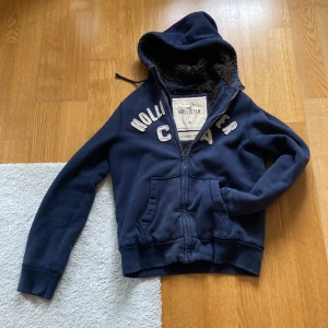 Mörkblå hoodie från Hollister med foder - Mysig mörkblå hoodie från Hollister med broderad text på bröstet. Tröjan har dragkedja, huva med snörning och fluffigt mörkbrunt foder på insidan. Två fickor framtill och ribbade muddar vid ärmslut och nederkant. Perfekt för kyliga dagar. Storlek M, har tecken på användning