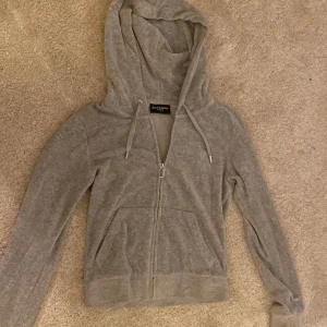 Grå Juicy Couture hoodie med dragkedja - Grå hoodie från Juicy Couture med huva, snörning och dragkedja med J-detalj. Mjuk och skön i bomullsmix, perfekt för chill dagar. Klassisk logga broderad på bröstet och två fickor framtill. Långärmad och normal passform.