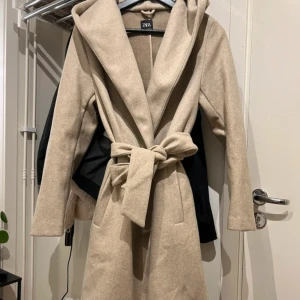 Beige kappa med huva från Zara - Stilren beige kappa från Zara med stor huva och knytskärp i midjan. Kappan har två svarta knappar framtill och raka, långa ärmar. Perfekt för dig som vill ha en klassisk look med modern twist.