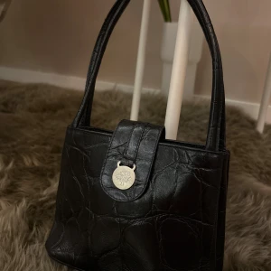 Svart handväska från Mulberry i skinn - Snygg svart handväska från Mulberry i croco-präglat skinn med silverfärgad logodetalj framtill. Väskan har två handtag och ett rutigt innerfoder med dragkedja. Perfekt storlek för det viktigaste och en klassisk look.