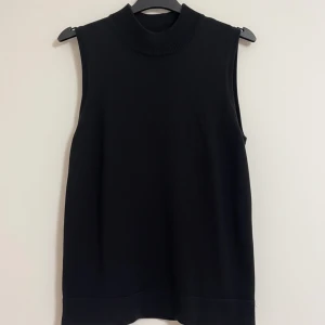 Svart stickad väst från KappAhl - Svart stickad väst från Number One by KappAhl med hög krage och rak passform. Västen är ärmlös och har en stilren, enkel design som funkar till många outfits. Perfekt att slänga över en skjorta eller t-shirt.