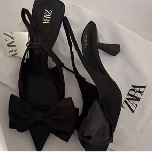 Svarta pumps med rosett från Zara - Stilrena svarta pumps från Zara med spetsig tå och stor dekorativ rosett framtill. Skorna har en elegant hög klack och smala remmar runt vristen. Perfekta för att lyfta din outfit med en trendig och chic touch.