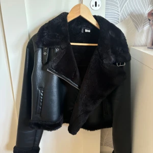 Svart pälsfodrad bikerjacka från H&M - Säljer en svart bikerinspirerad jacka från H&M med mjukt fuskpälsfoder och stora slag. Jackan har dragkedja, detaljer i silver och är croppad i modellen. Perfekt för dig som vill ha en edgy look med extra värme.