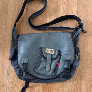 Grå axelväska från Diesel - Snygg grå axelväska från Diesel med justerbar rem och flera praktiska, stora fack. Väskan har detaljer i svart och metall, samt en framficka med Diesel-logga. Köpt på humana men inte använt den 💗💗