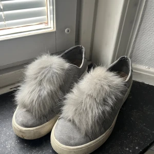 Grå sneakers med fuskpäls från Steve Madden - Grå sneakers från Steve Madden med fluffig fuskpäls på ovansidan. Skorna har en platt vit sula och är tillverkade i mockaliknande material. Perfekt för dig som vill sticka ut med en unik och trendig detalj på fötterna.