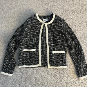 Svartvit cardigan jacka från Kappahl - Stilren svartvit cardigan jacka från Kappahl med klassiskt rutigt mönster och kontrasterande vita kanter. Jackan har två fickor framtill och är långärmad. 