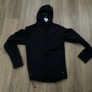 Svart vindjacka från Cross Sportswear - Svart vindjacka med huva från Cross Sportswear. Jackan har hel dragkedja framtill, två fickor med dragkedja och justerbar nederkant. Enkel och stilren design, perfekt för blåsiga dagar. Materialet är mjukt och smidigt syntet.