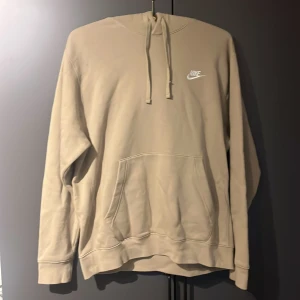 Beige Nike hoodie med huva - Säljer en beige hoodie från Nike med klassisk logga på bröstet. Tröjan har huva med snörning, stor magficka och långa ärmar. Perfekt för chill dagar eller när du vill ha en clean och enkel look.