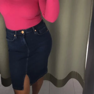 Mörkblå jeanskjol med slits - Snygg mörkblå jeanskjol i klassisk denim med hög midja och slits framtill. Kjolen har fickor både fram och bak samt gylf med knapp. Perfekt för dig som gillar en stilren och trendig look.