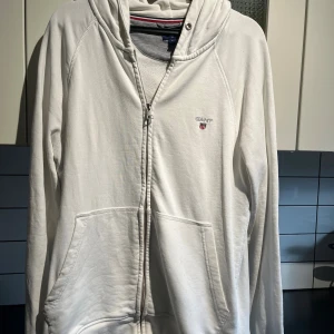 Vit hoodie med dragkedja från Gant - Vit hoodie från Gant med dragkedja framtill och klassisk huva. Tröjan har två stora fickor fram och Gant-logga broderad på bröstet. Skön passform och perfekt för lager-på-lager. Snygg och enkel stil som funkar året runt.
