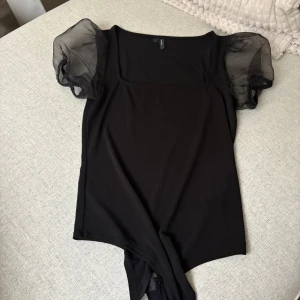 Svart body med puffärmar från Vero Moda - Svart body från Vero Moda med transparenta puffärmar i mesh. Toppen har en fyrkantig urringning och är figurnära med korta ärmar. Perfekt för dig som gillar en stilren look med en twist. Materialet är mjukt och stretchigt, vilket gör den bekväm att bära.