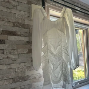 Vit kort klänning från NLY Trend - Supersöt vit kort klänning från NLY Trend med långa puffärmar och snygg rynkad effekt framtill. Klänningen har dragkedja i sidan och är gjord i ett tunt, lätt material som ger en luftig känsla. Perfekt för dig som vill sticka ut med en romantisk look.