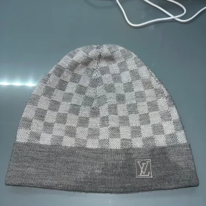 Grå rutig Louis Vuitton mössa - Snygg grå och vit mössa från Louis Vuitton med klassiskt rutigt mönster och LV-logga framtill. Mössan är stickad i mjukt material och har en bred uppvikt kant. Perfekt för dig som vill ha en lyxig och stilren accessoar till vintern. Den är ny tar byten beror på vad det är pris kan diskuteras 
