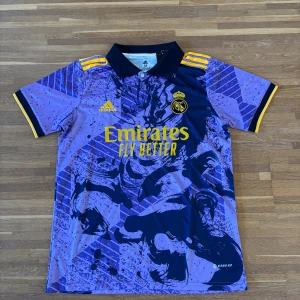 Real Madrid lila bortatröja Adidas M  | Lätt & ventilerande - Säljer en Real Madrid bortatröja från Adidas i storlek M, i lila och mörkblått med gula detaljer. Tröjan har korta ärmar, svart krage och tryckt klubbmärke samt sponsorlogga. Tillverkad i lätt och ventilerande material – perfekt för fotboll, träning eller streetwear.  ✔️ Lätt och andas ✔️ Kortärmad, regular fit ✔️ Aldrig använd  Storlek: M Skick: Ny / aldrig använd 