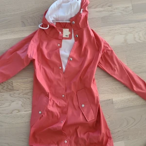 Koralröd regnjacka från 157, XS - Snygg korallröd regnjacka från 157 med huva och vita detaljer. Jackan har tryckknappar, två stora fickor framtill och är helfodrad i vitt. Perfekt för regniga dagar och har en clean, stilren look.