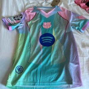 Barça Lamine Yamal Nike fotbollströja - Säljer en limited edition FC Barcelona fotbollströja från Nike med Lamine Yamal och nummer 19 på ryggen. Tröjan har korta ärmar, coolt mönster i pastellfärger som blå, rosa, grön och gul, samt Champions League-märke på ärmen. Materialet är lätt och andas, perfekt för match eller träning.