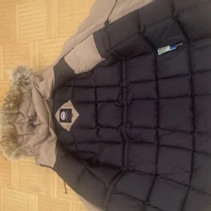 Beige och svart dunjacka Canada Goose M - Säljer en Canada Goose dunjacka i beige och svart med pälskantad huva. Jackan är quiltad och har klassisk logga på insidan. Perfekt för kalla vinterdagar och har en riktigt snygg streetstil.