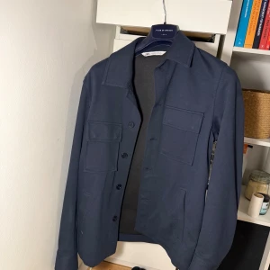Mörkblå overshirt från Zara - Stilren mörkblå overshirt från Zara med klassisk krage, fyra fickor framtill och knäppning med svarta knappar. Perfekt lager-på-lager-plagg med en clean look och raka linjer. Snygg att bära öppen eller stängd.