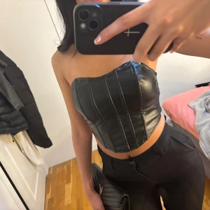 Svart korsett-topp i skinnimitation - Säljer en svart korsett-topp i skinnimitation med markerade sömmar och dragkedja bak. Toppen är croppad och axelbandslös, perfekt för att skapa en edgy look. Snygg passform som framhäver midjan.