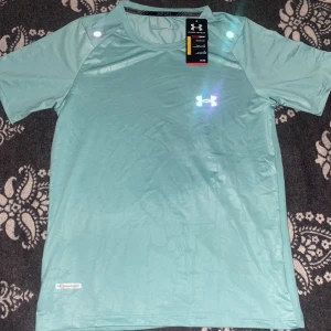 Under Armour reflective turkos t-shirt - Snygg turkos t-shirt från Under Armour med HeatGear-teknologi som håller dig sval och torr. Perfekt för träning, med reflekterande logga på bröstet och lätt, snabbtorkande material. Klassisk rund hals och korta ärmar för maximal rörelsefrihet.