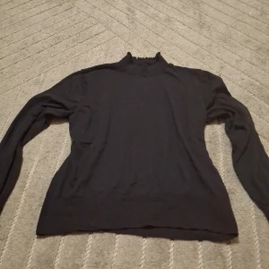 Svart polotröja med volang från H&M - Svart polotröja från H&M med lång ärm och volangdetaljer vid hals och ärmslut. Tröjan är stickad i mjukt material och har en klassisk, enkel look med en liten twist tack vare volangerna. Perfekt för lager-på-lager under kyliga dagar.