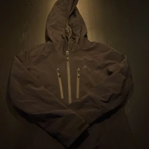 Svart vindjacka från Five Seasons - Svart vindjacka med huva från Five Seasons. Jackan har två vertikala bröstfickor med dragkedjor och en diskret logga på bröstet. Materialet är syntet och jackan har långa ärmar samt justerbara muddar. Perfekt för blåsiga dagar.