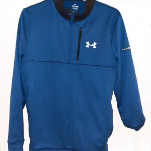 Blå half zip Under Armour hoodie - Blå hoodie från Under Armour med half zip och svart detalj vid dragkedjan. Tröjan har lång ärm, vit logga på bröstet och är gjord i ett mjukt, sportigt material som passar perfekt till träning. Snygg och enkel design med normal passform.