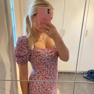 Blommig långklänning från JDY - Superfin långklänning från JDY med blommigt mönster i rosa, blått och rött. Klänningen har puffärmar, söt v-ringning och en slits på sidan. Perfekt för sommarens alla tillfällen. Materialet är mjukt och lätt, troligtvis polyester.