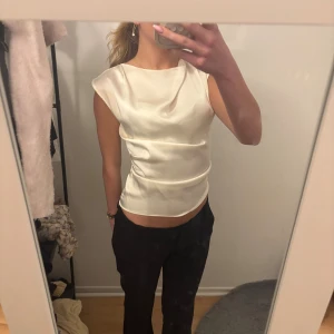 Vit satintopp med ärmlös design - Säljer en stilren vit topp i satin med ärmlös design och lätt glansig finish. Toppen har en rak, något lös passform och rundad halsringning. Perfekt för dig som gillar en clean och elegant look.