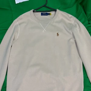 Beige långärmad tröja Polo Ralph Lauren - Snygg beige långärmad tröja från Polo Ralph Lauren med klassisk rund halsringning och broderad logga på bröstet. Tillverkad i mjuk bomull som känns skön mot huden. Perfekt för lager-på-lager och enkel att matcha med jeans eller chinos.Liknande modell som originalet!
