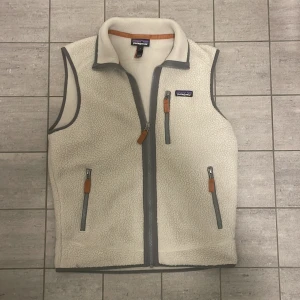 Vit fleeceväst från Patagonia S - Mysig vit fleeceväst från Patagonia i storlek S. Västen har grå detaljer, hög krage och tre fickor med dragkedja och orangea band. Perfekt för lager-på-lager och outdoor-vibes. Klassisk Patagonia-logga på bröstet.