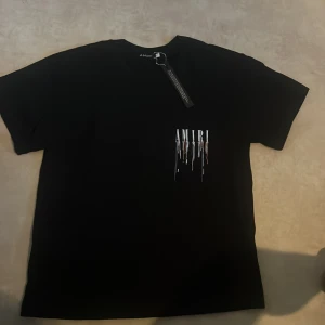 Svart AMIRI t-shirt med färgstänk - Svart t-shirt från AMIRI i bomull med stort tryck på ryggen och mindre tryck på bröstet. AMIRI-loggan har coola färgstänk i vitt, lila, orange och rött som rinner ner. Klassisk passform och korta ärmar, perfekt för en streetwear-look.