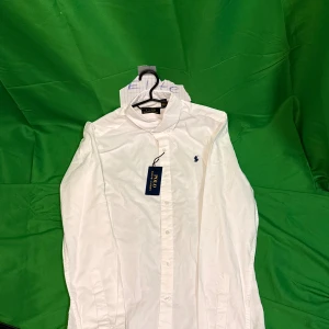 Vit skjorta från Polo Ralph Lauren - Klassisk vit skjorta från Polo Ralph Lauren med button down-krage och broderad marinblå logga på bröstet. Skjortan är långärmad, har knappar framtill och är tillverkad i mjuk bomull. Perfekt för en clean och stilren look.Liknande modell som originalet!
