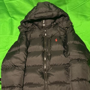 Svart pufferjacka från Polo Ralph Lauren - Svart pufferjacka från Polo Ralph Lauren med röd broderad logga på bröstet. Jackan har huva, dragkedja framtill och flera fickor. Perfekt för kalla dagar och riktigt snygg streetstil.Liknande modell som originalet!