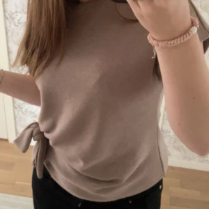 Beige topp med knyt i sidan - Säljer en beige topp med snygg knytning i sidan. Toppen är ärmlös och har en enkel, stilren design som passar perfekt till jeans eller kjol. Mjuk och skön att bära, perfekt för vår och sommar.
