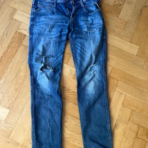 Blåa replays - Säljer ett par blå Replay Anbass jeans med snygga slitningar och hål på benen. Klassisk femficksmodell med detaljerade sömmar på bakfickan och Replay-logga vid fickan. Jeansen har en normal passform och är tillverkade i mjukt denimtyg.