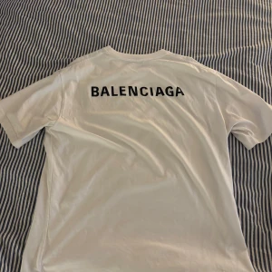 Vit Balenciaga t-shirt unisex - Vit t-shirt från Balenciaga med svart logga tryckt på bröstet och stort på ryggen. Unisex-modell med klassisk passform och rund hals. Tillverkad i mjuk bomull för en clean och stilren look. T-shirten är givetvis äkta och köpt på miinto.se ganska oversized (se bild) är 188cm 86kg