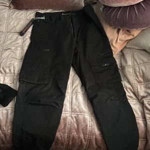 Svarta cargopants från Texstar 30/30 ny med prislapp på - Svarta cargopants från Texstar med flera praktiska fickor på benen och klassisk cargomodell. Byxorna har rak passform och är tillverkade i slitstarkt material, perfekta för dig som gillar funktionella och stilrena byxor.