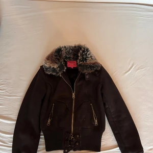 Brun mocka bomberjacka med fuskpäls - Snygg mörkbrun bomberjacka från Portobello Princess i mocka med fluffig fuskpäls runt kragen. Jackan har dragkedja framtill, två snedställda fickor med dragkedjor och ribbade muddar nertill. Insidan är fodrad med mjuk pälsimitation för extra värme.