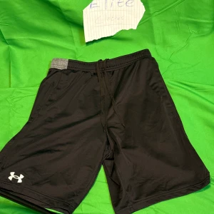 Svarta träningsshorts från Under Armour - Svarta träningsshorts från Under Armour med grå resår i midjan och snörning framtill. Shortsen har en loose passform och är tillverkade i ett lätt syntetmaterial som andas. Perfekta för gymmet eller löprundan, med diskret logga på benet.Liknande modell som originalet!
