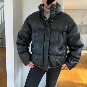 Svart pufferjacka i skinnimitation - Säljer en svart pufferjacka med glansig finish i skinnimitation. Märke: Divided H&M. Jackan har hög krage, dragkedja och tryckknappar framtill samt stora fickor. Oversized passform som ger en cool och trendig look! Stl S