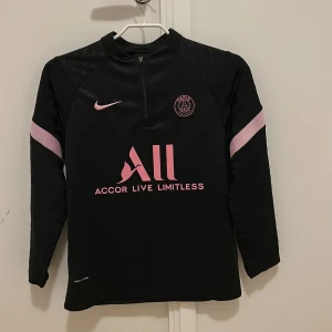 Svart PSG långärmad tröja  - Säljer en svart långärmad Paris Saint-Germain tränings tröja från Nike med rosa detaljer och logga. Tröjan har en dragkedja framtill och är i storlek 158 cm. Hör av er vid frågor!