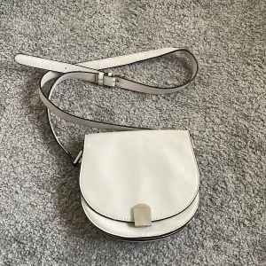Vit axelväska i skinn - Snygg vit axelväska i skinn med rundad form och justerbart axelband. Väskan har ett stilrent silverfärgat spänne framtill och minimalistisk design som passar till många outfits.