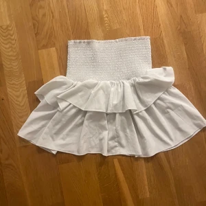 Vit volangkjol med smock - Supersöt vit kortkjol med bred smock i midjan och dubbla volanger. Kjolen har en luftig och flowy känsla, perfekt för varma dagar. Enkel att matcha med crop tops eller linnen för en fräsch look.