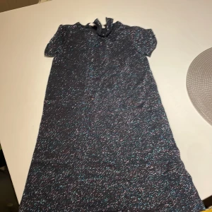 Glittrig mörkblå klänning H&M 122/128 - Supersöt mörkblå glittrig klänning från H&M i storlek 122/128. Klänningen har korta ärmar och en rak, enkel passform. Perfekt för dig som gillar glitter och vill sticka ut lite extra. Snygg detalj med liten rosett i halsen.